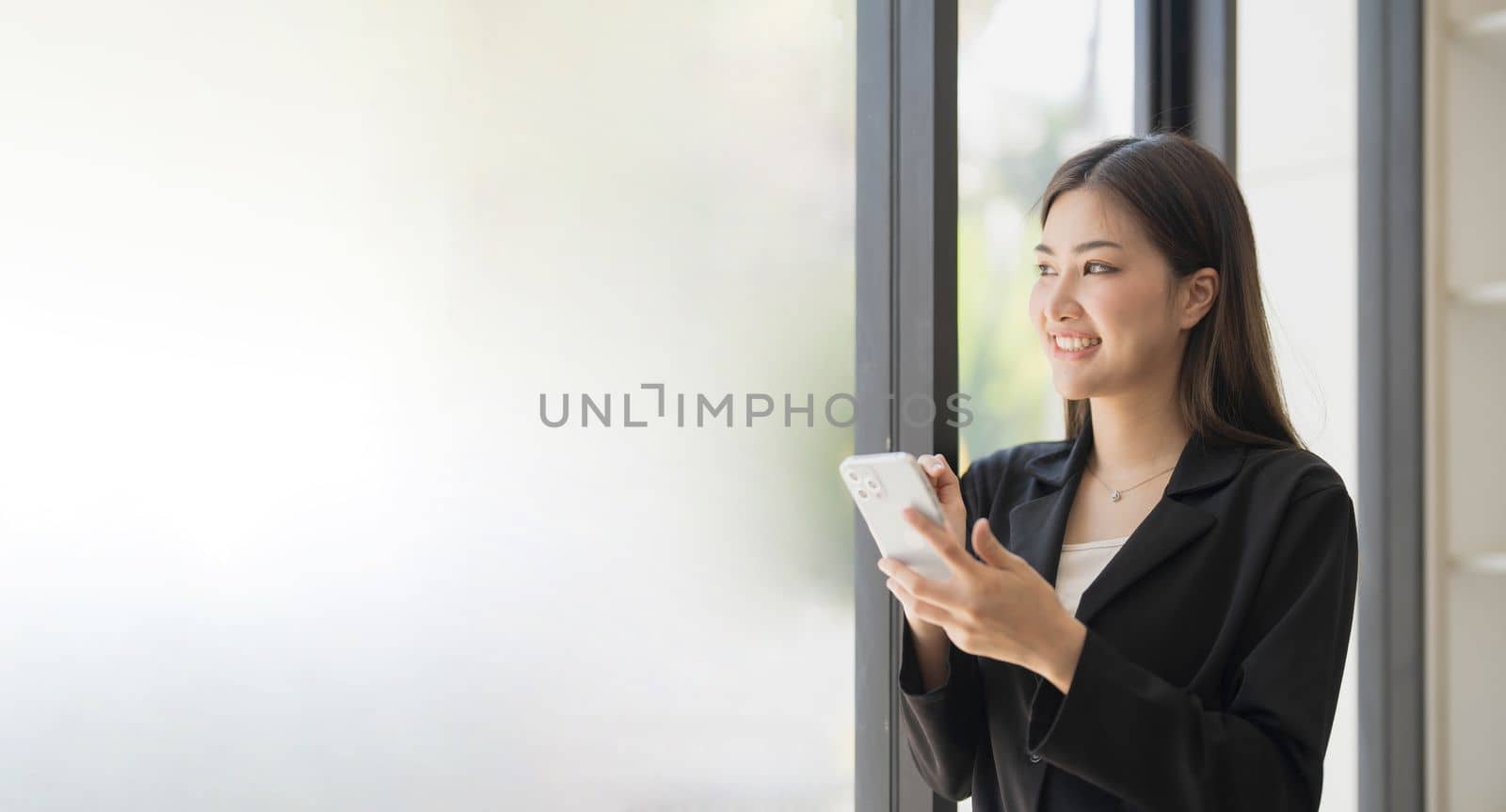 Portrait of elegant successful asian business woman speaking by smartphone and smiling happily while standing against window in office