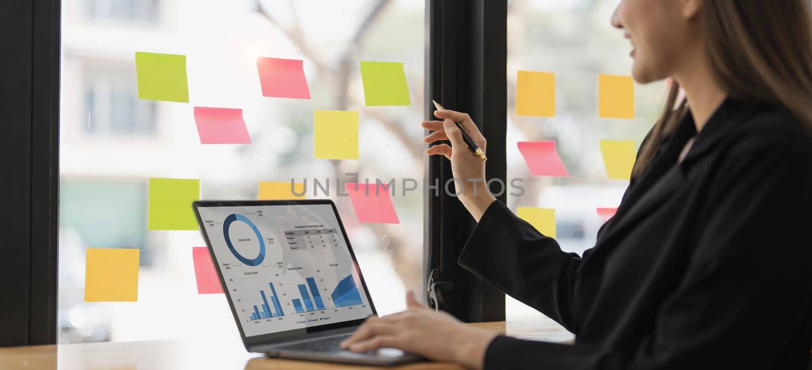 Young smiley attractive, businesswoman using laptop and post it notes in glass wall to writing strategy business plan to development grow to success by wichayada