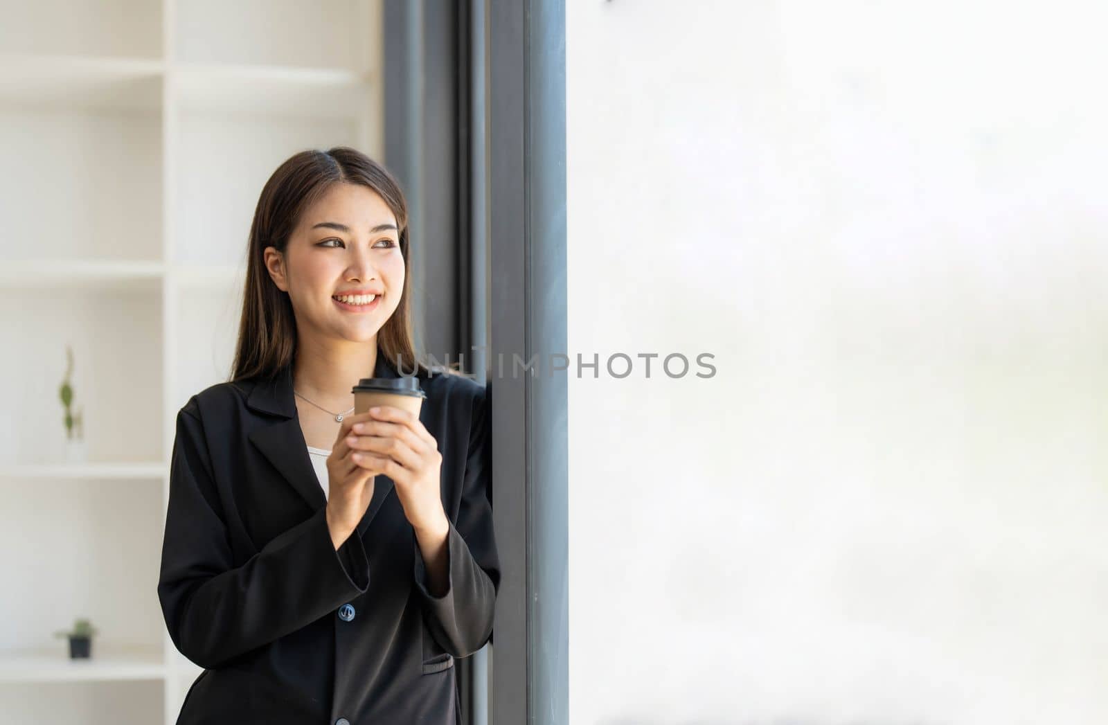 Portrait of young confident woman holding coffee cup, standing at office and looking outside. Successful businesswoman standing in office with copy space. by wichayada