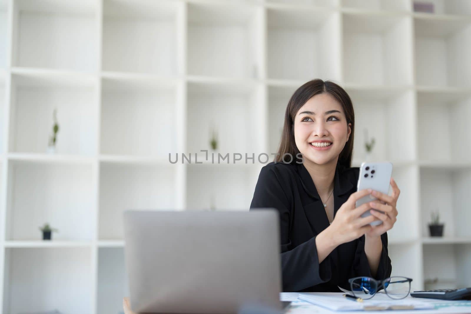 Smiling beautiful Asian businesswoman analyzing chart and graph showing changes on the market and holding smartphone at office. by wichayada