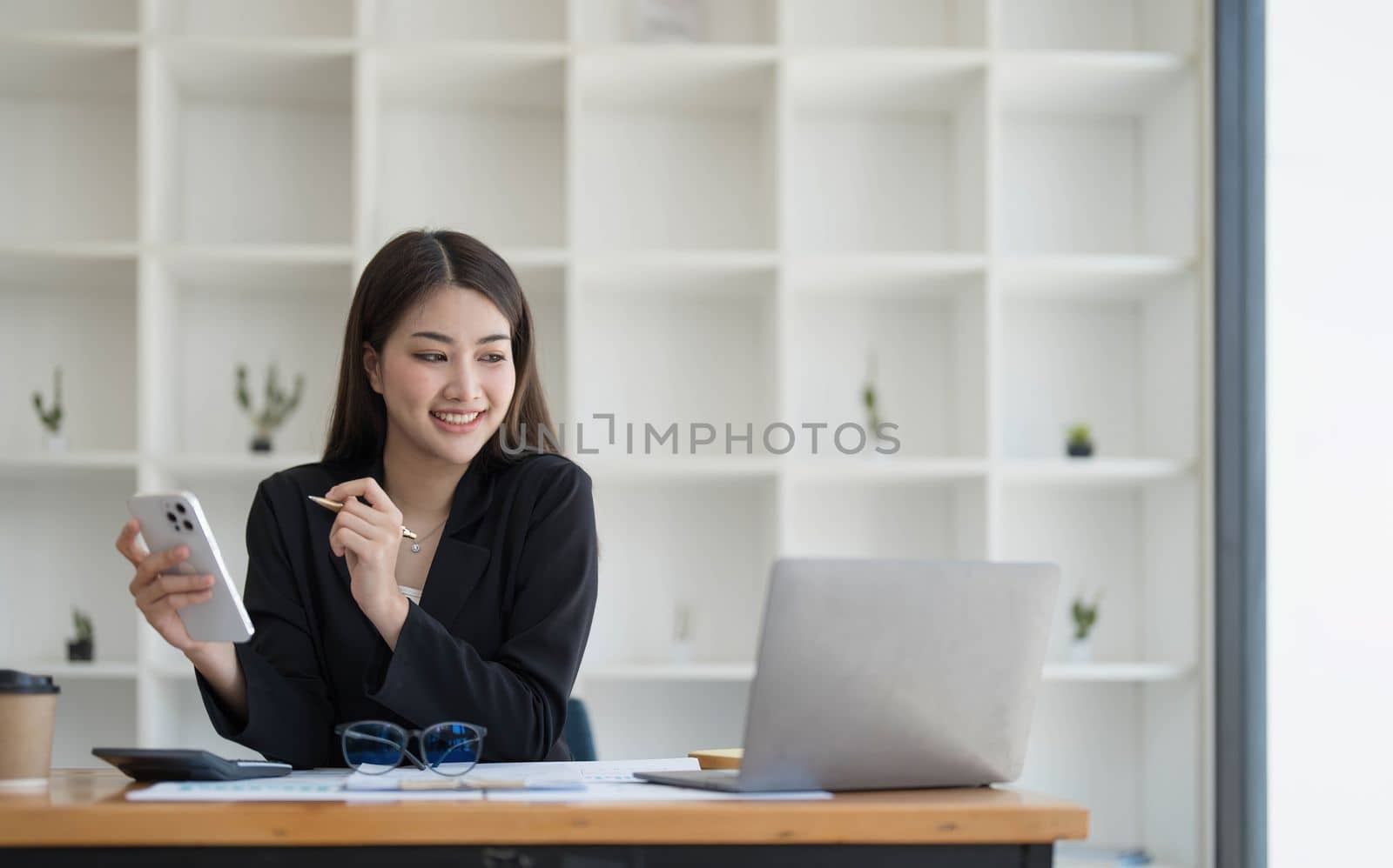 Smiling beautiful Asian businesswoman analyzing chart and graph showing changes on the market and holding smartphone at office. by wichayada