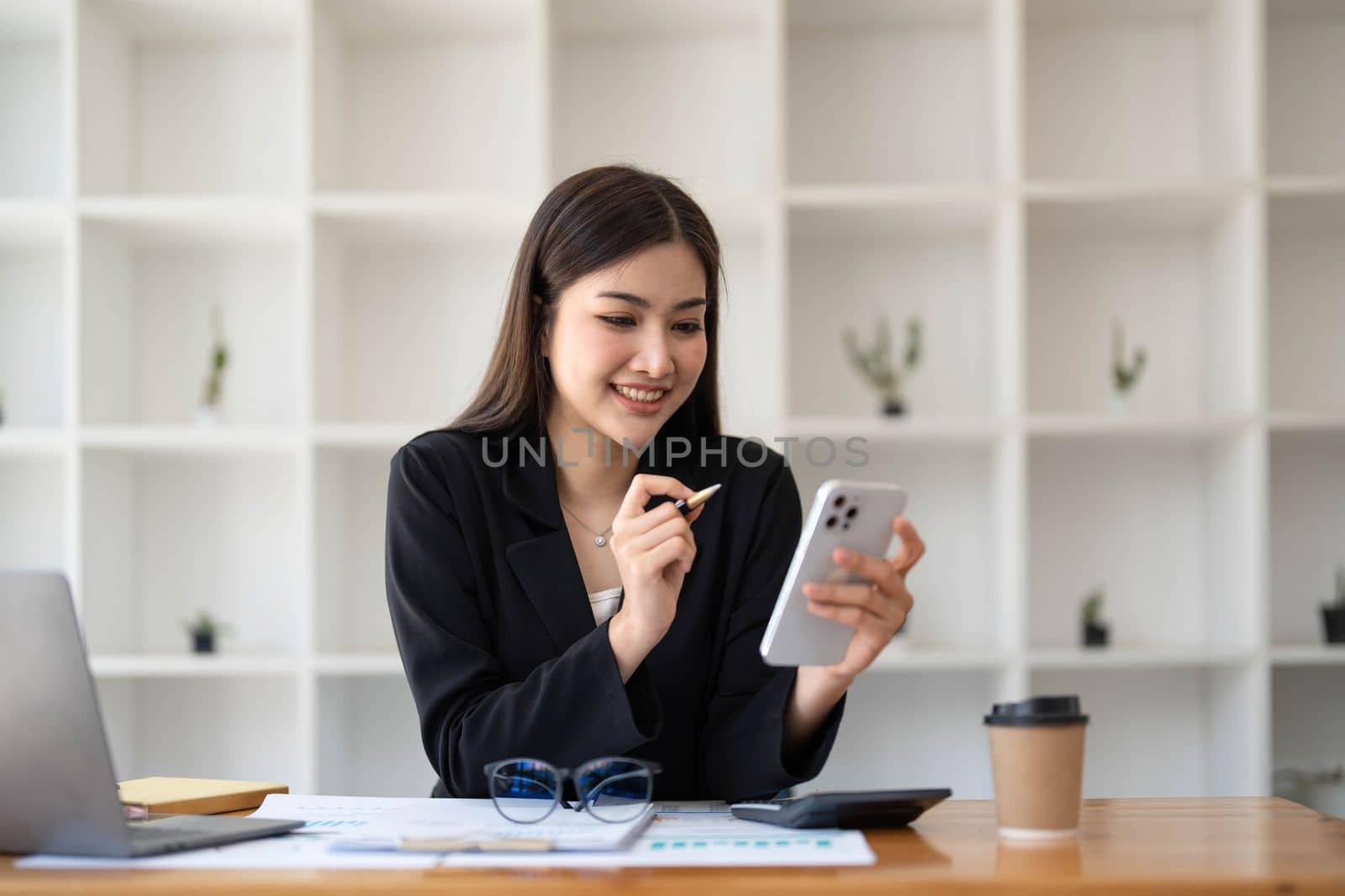 Smiling beautiful Asian businesswoman analyzing chart and graph showing changes on the market and holding smartphone at office. by wichayada