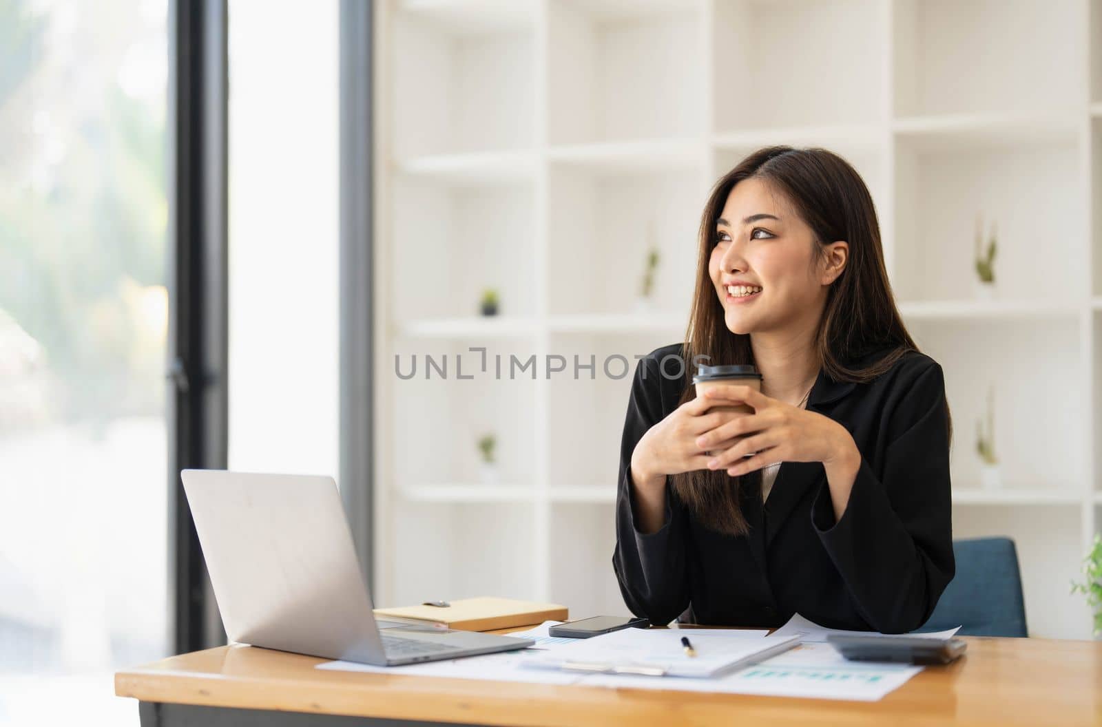Beautiful young Asian businesswoman smiling holding a coffee mug and laptop working at the office. by wichayada