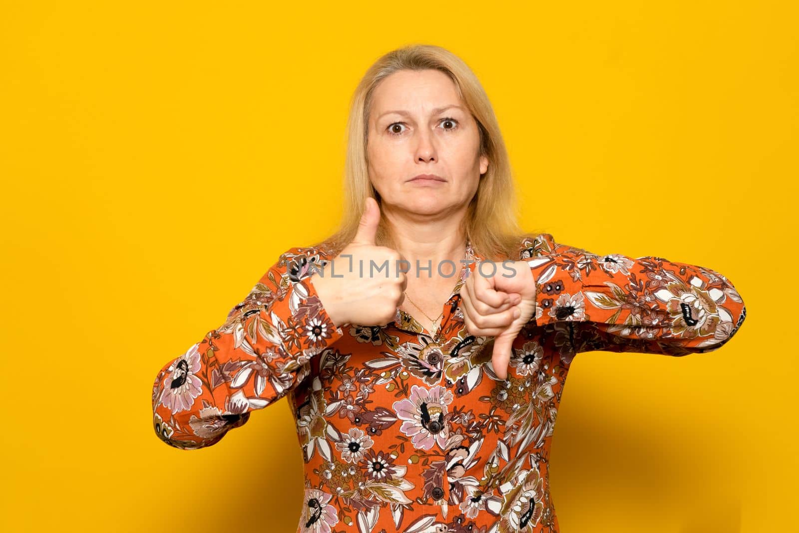 Pretty caucasian blonde woman in a patterned dress giving one thumbs up and one thumbs down isolated over yellow background. doubt concept