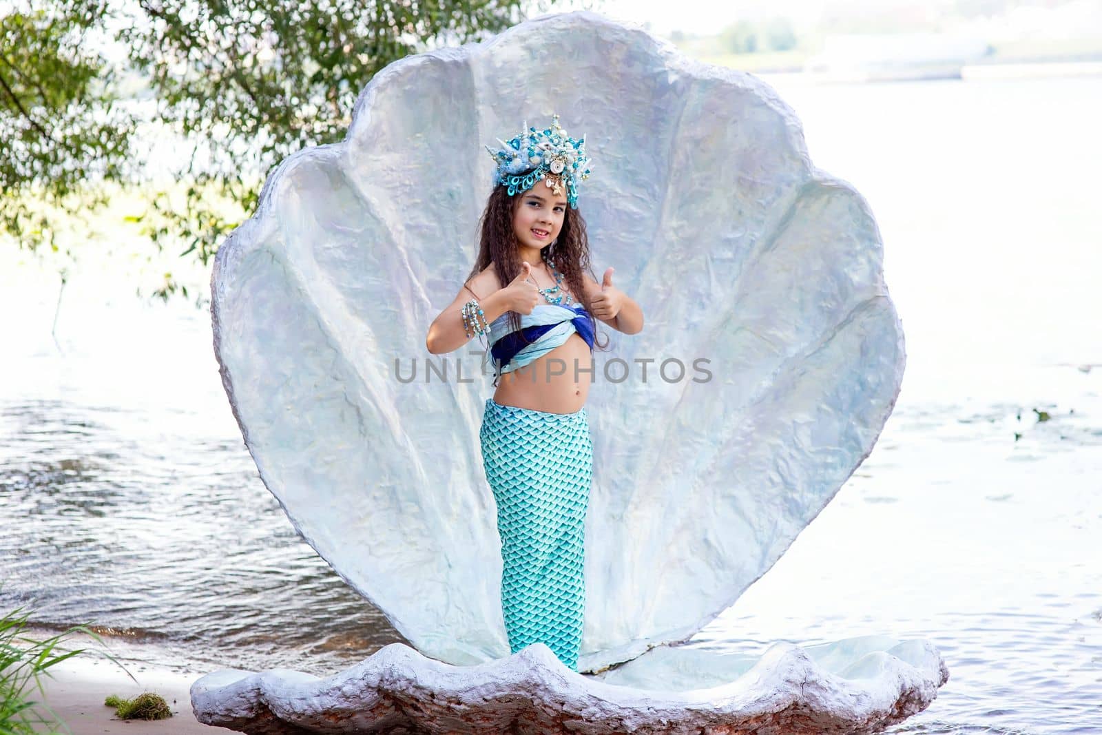 A cute little girl in a mermaid costume stands outdoor, in a large sea shell, holds her thumbs up. Copy space