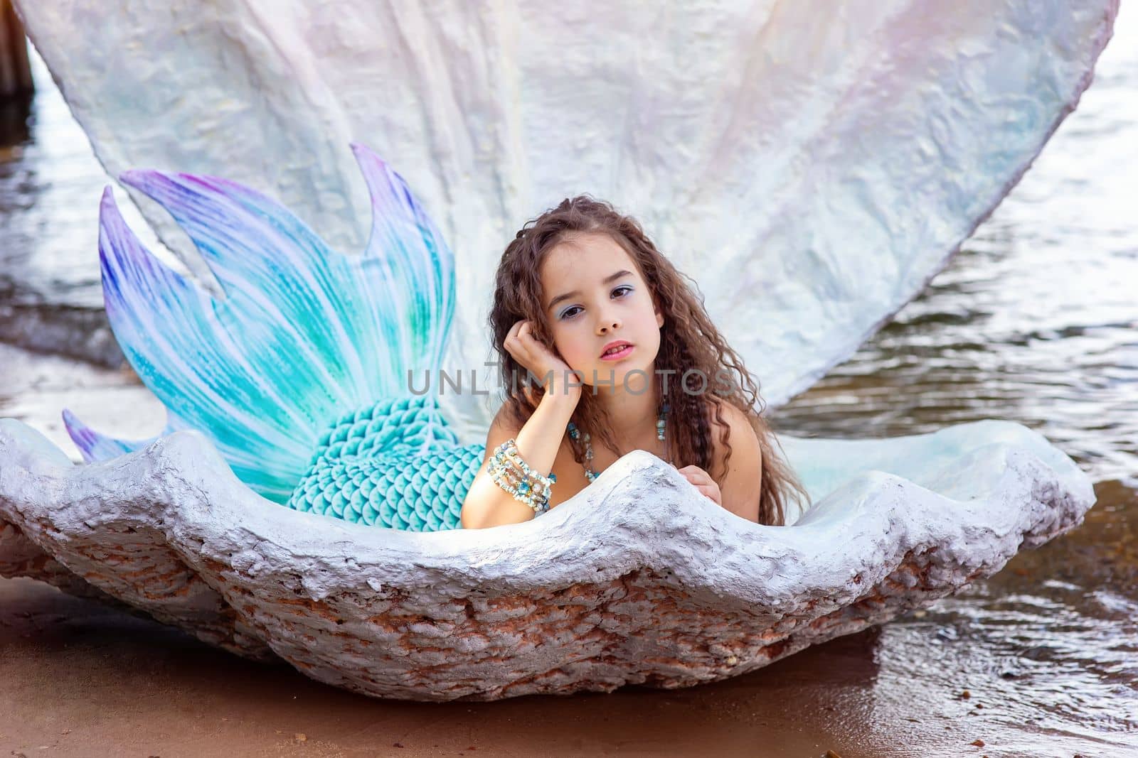 A pretty little brunette girl in a turquoise mermaid costume lie in a large sea shell, outdoor. Close up. Copy space