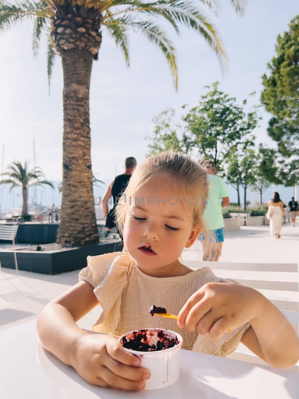 Little girl sits at a table on the street and eats popsicles with a spatula from a cup by Nadtochiy