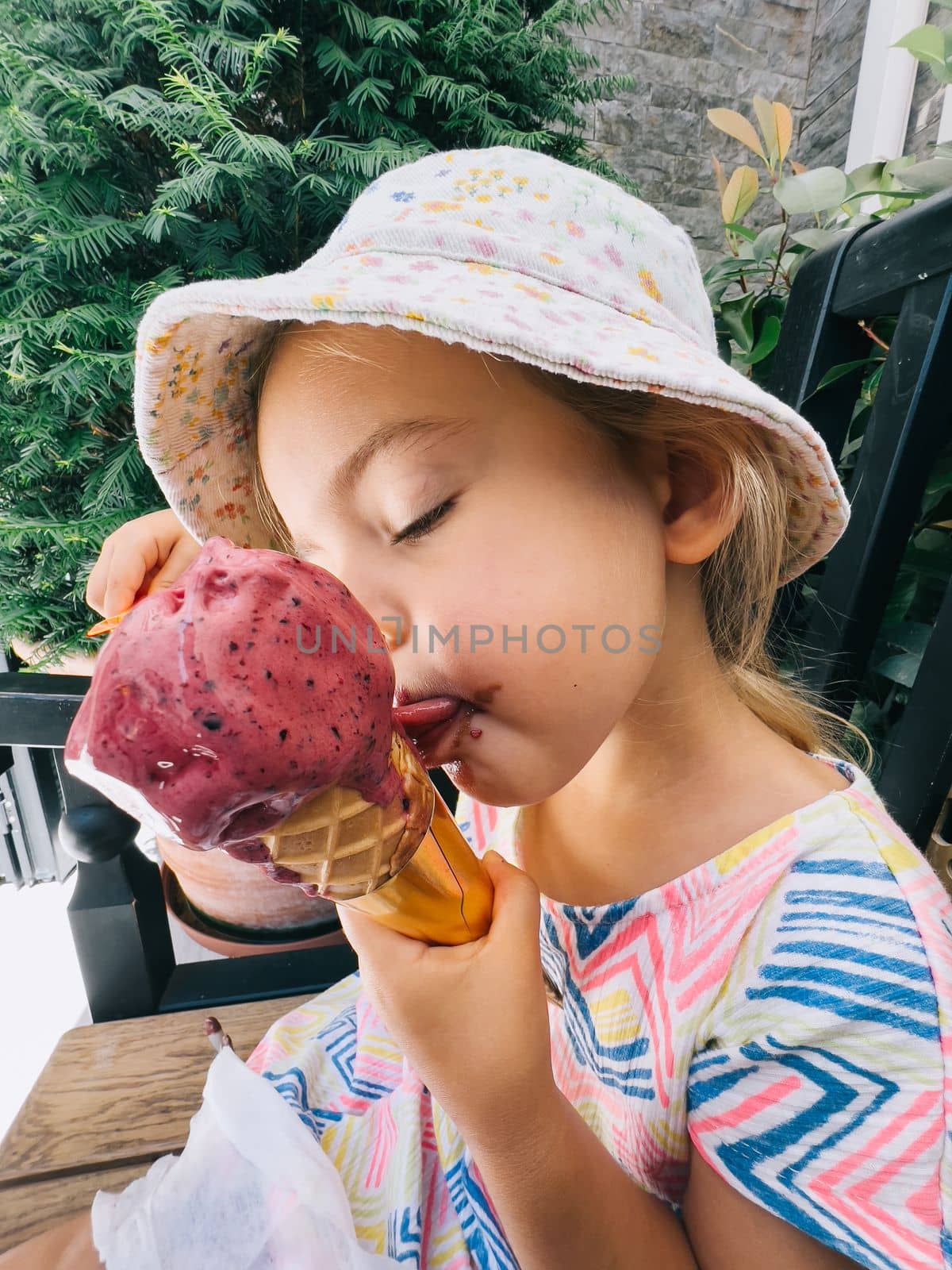 Little girl licks a popsicle in a cone sitting on a bench. High quality photo