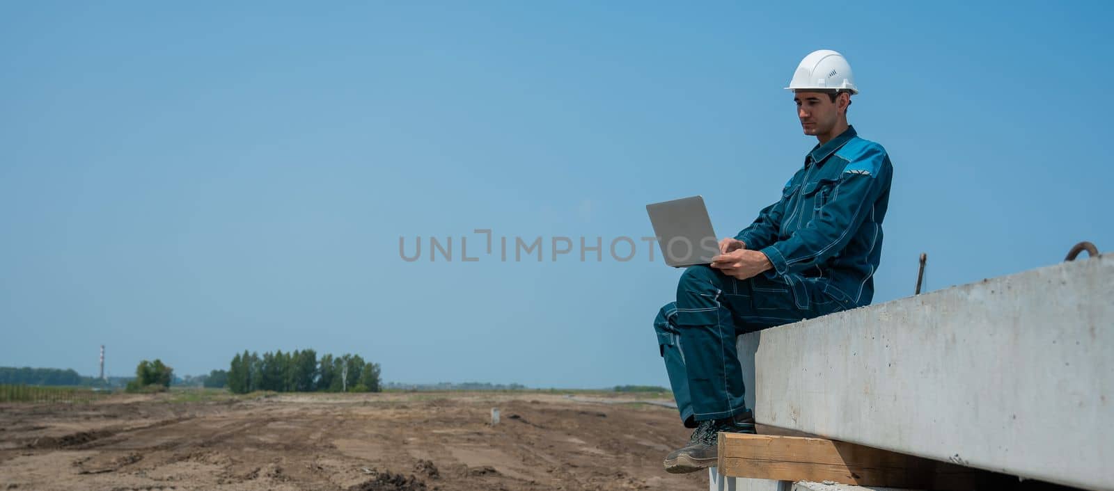 Caucasian male builder in hardhat sits on floor slabs and uses laptop at construction site. by mrwed54