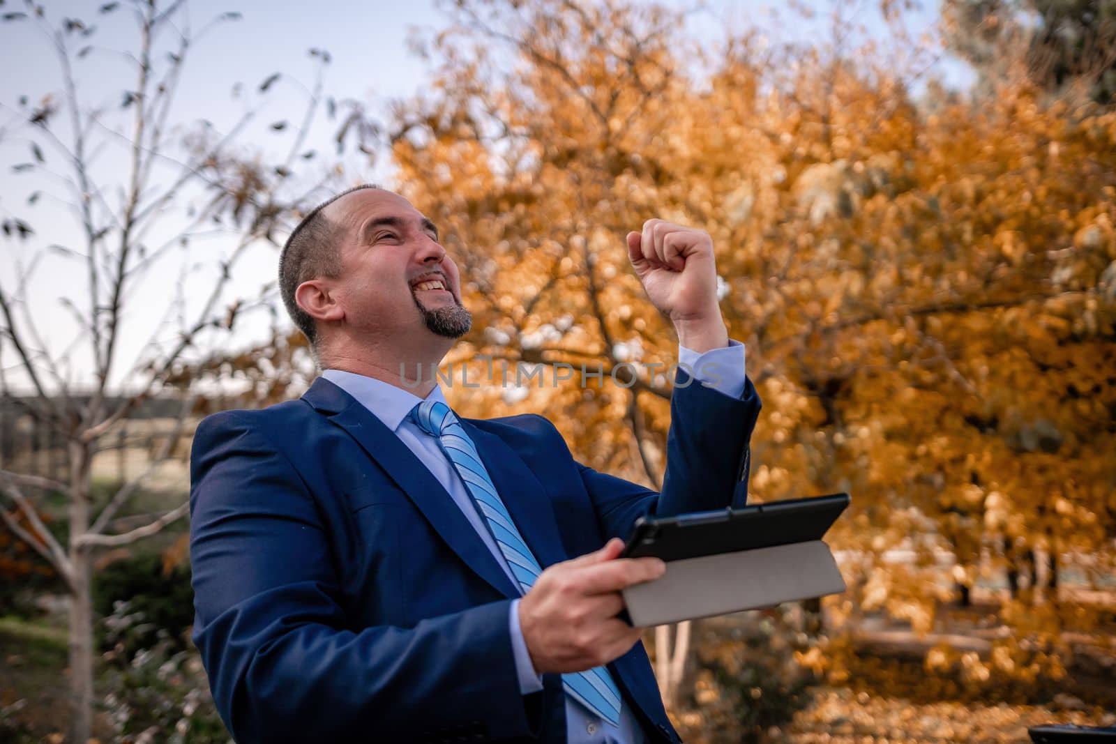 Mature business man in suit reading a news sitting in the park on the bench. Entrepreneur drink coffee in autumn park. Senior executive rest in city park. by panophotograph