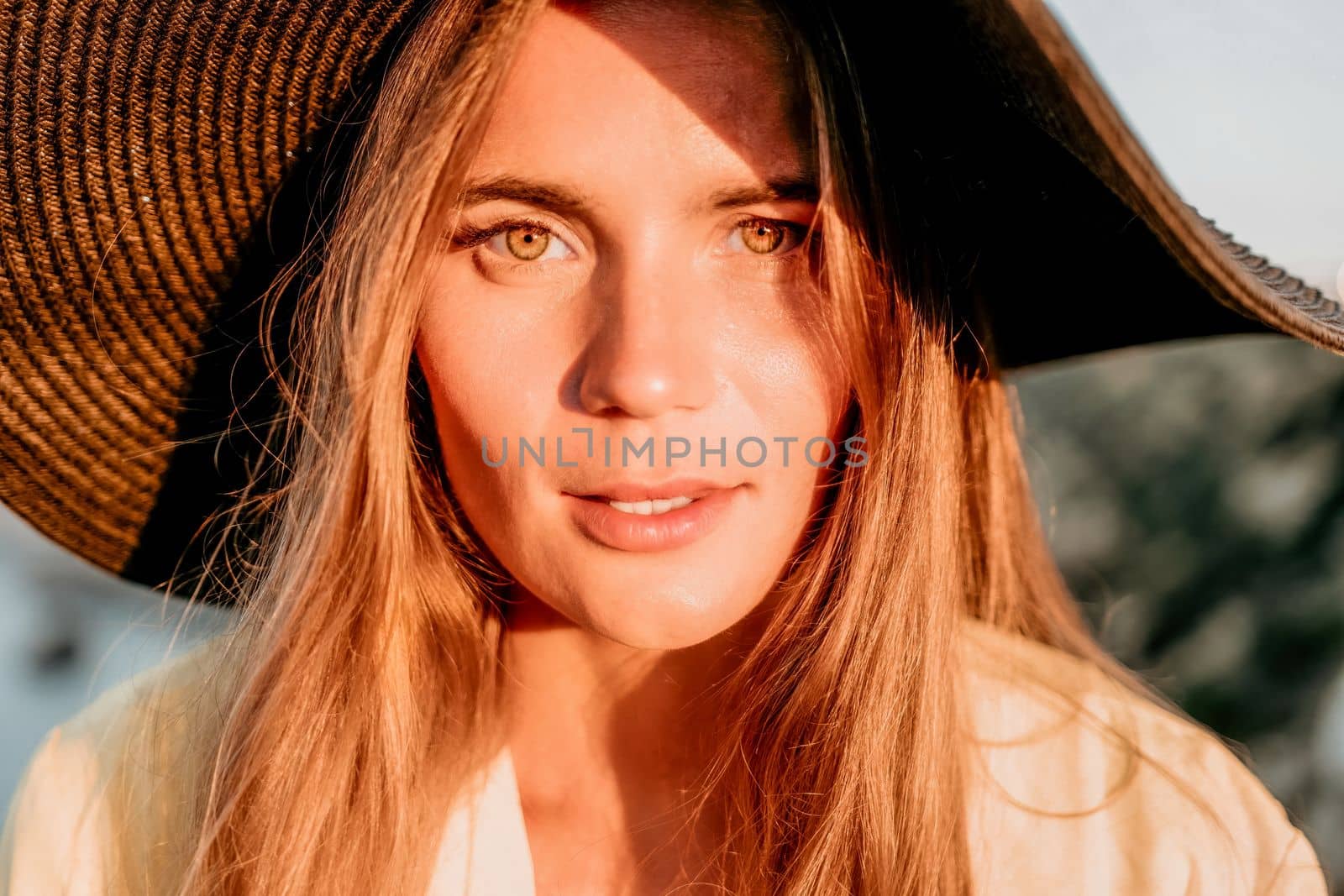 Portrait of happy young woman wearing summer black hat with large brim at beach on sunset. Closeup face of attractive girl with black straw hat. Happy young woman smiling and looking at camera at sea