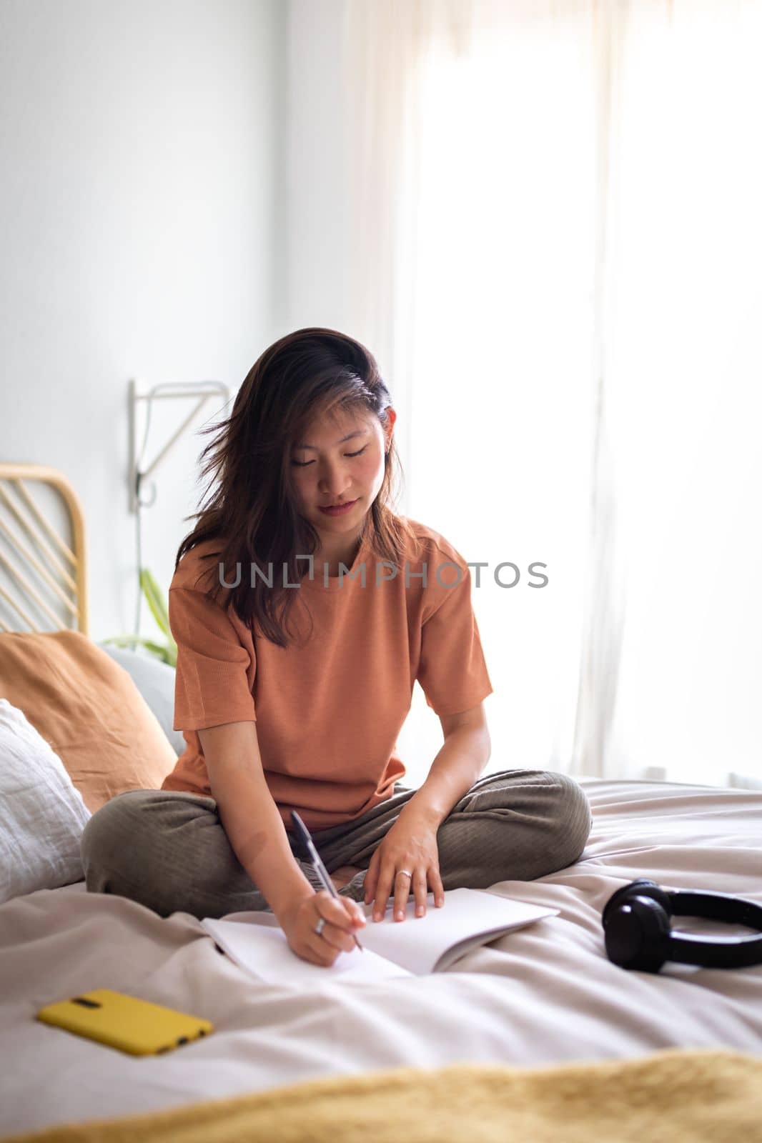 Asian female teen college student sitting on bed writing on journal in cozy bedroom. Copy space. Vertical image. Lifestyle concept.