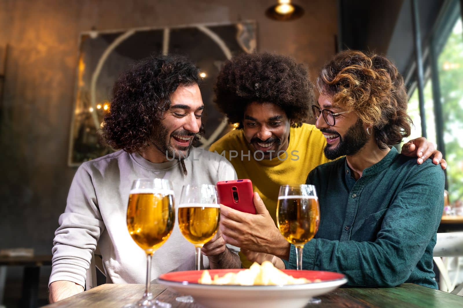 Young man showing to male friends mobile phone while having drinks together in pub.Technology and leisure activity.