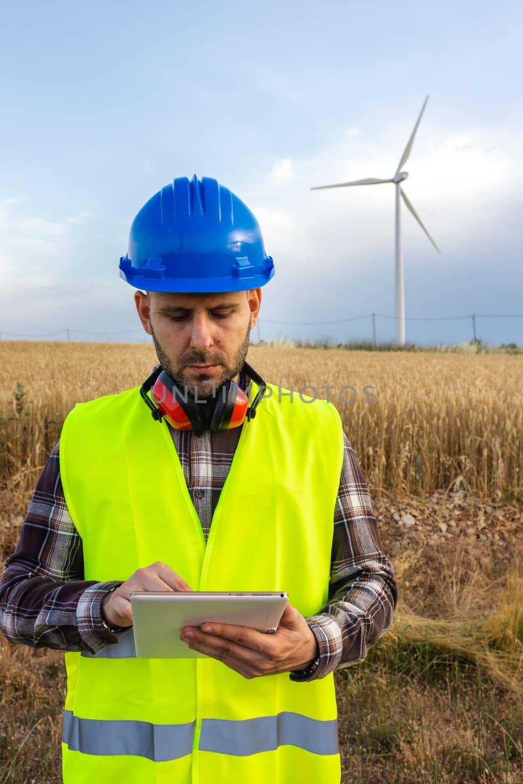 Male wind turbine farm maintenance worker wearing helmet and vest checking data on digital tablet. Vertical image. Renewable energies concept.