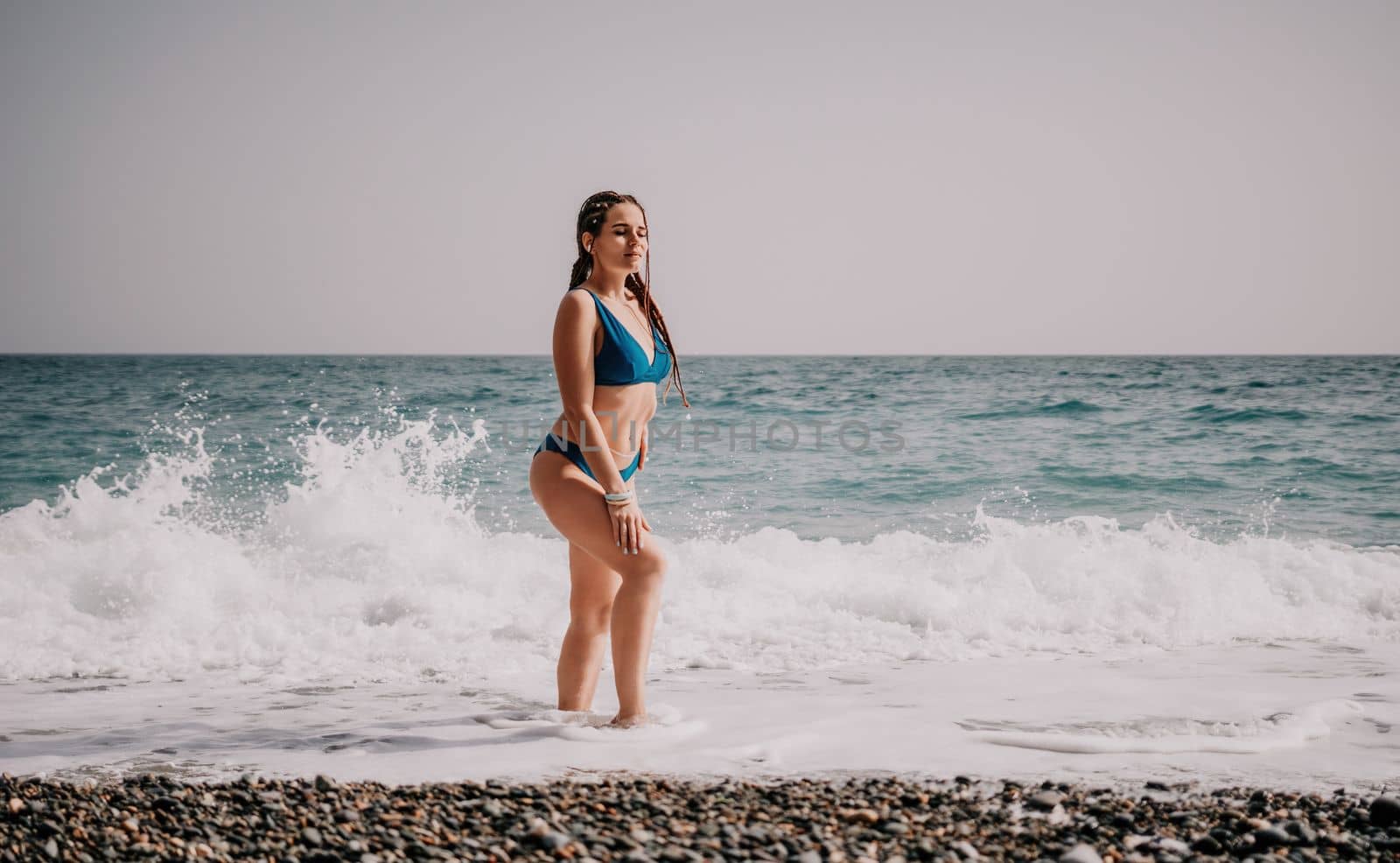 A tourist woman in blue bikini on beach enjoys the turquoise sea during her summer holiday. Rear view. Beach vacation. woman with braids dreadlocks standing with her arms raised enjoying beach ocean. by panophotograph