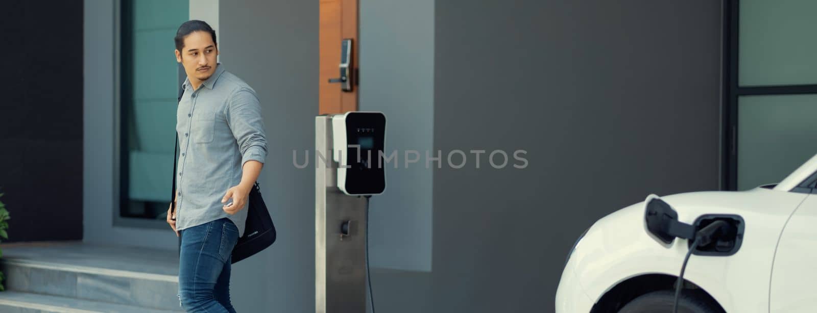 Progressive asian man and electric car with home charging station. Concept of the use of electric vehicles in a progressive lifestyle contributes to a clean and healthy environment.