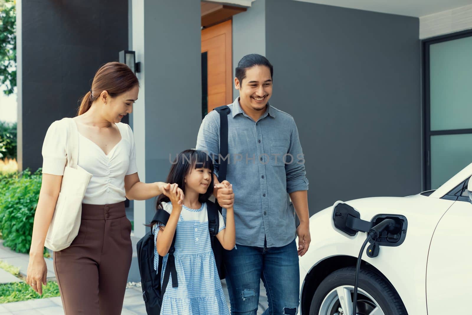 Progressive young parents and daughter with electric vehicle and home charging station. Green and clean energy from electric vehicles for healthy environment. Eco power from renewable source at home.