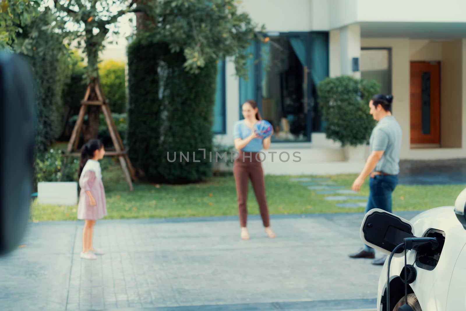 Focus closeup electric vehicle recharging battery from home electric charging station with blurred family in background. Renewable clean energy car for progressive eco awareness lifestyle concept.