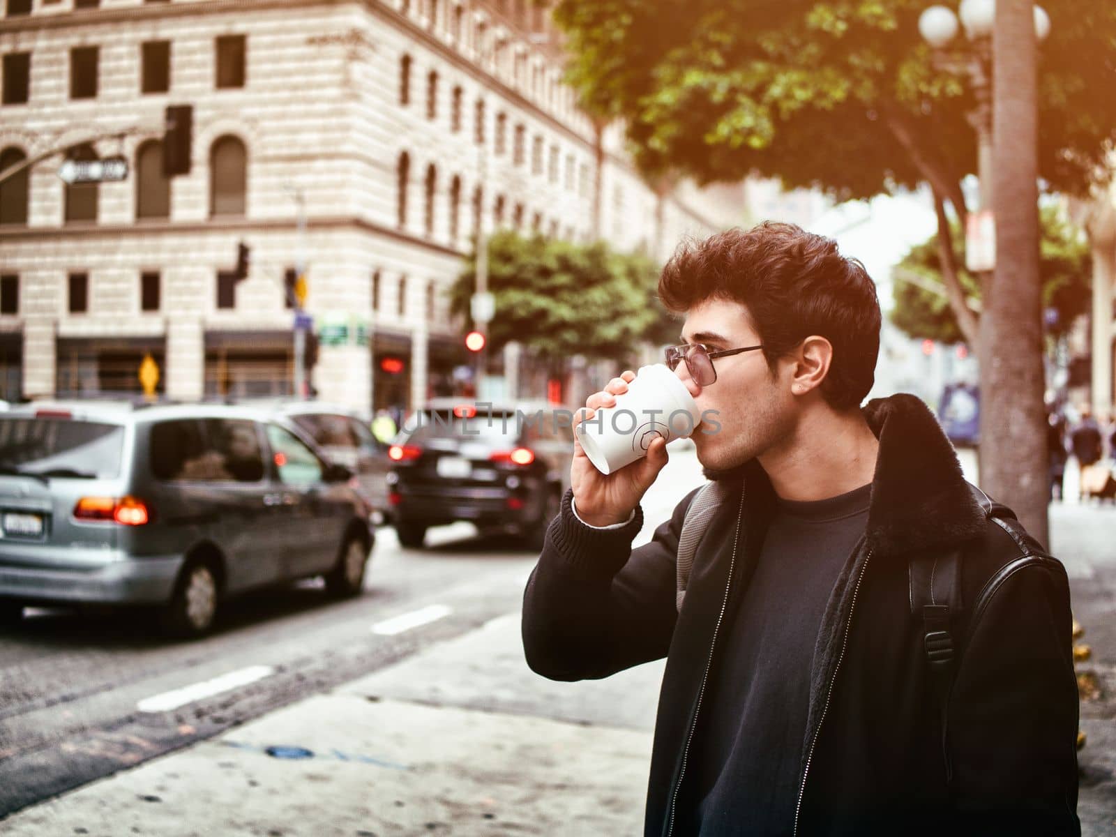 Attractive young man in modern city center drinking coffee from paper cup, wearing leather jacket