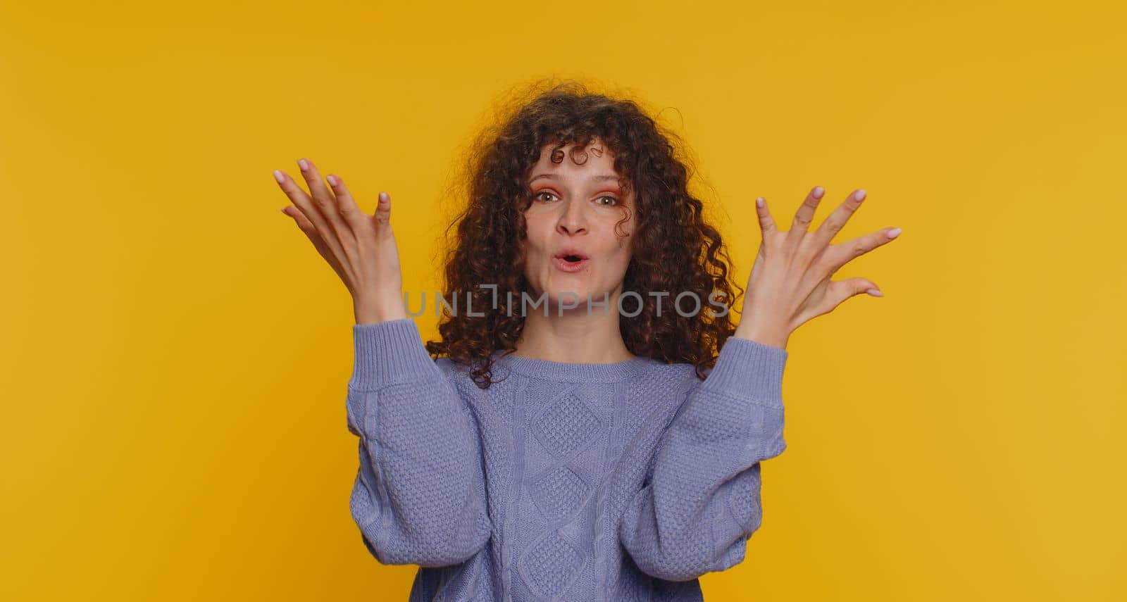 Oh my God, Wow. Excited amazed winner woman raising hands in surprise looking at camera with big eyes, shocked by sudden victory. Sincere young curly haired girl isolated on yellow studio background