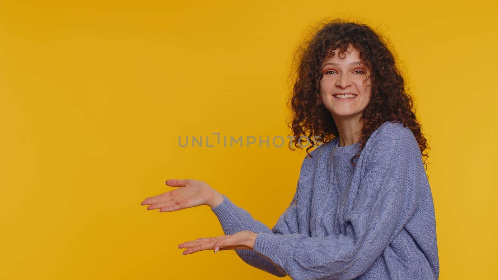 Lovely happy curly haired woman showing thumbs up and pointing empty place, advertising area for commercial text, copy space for goods promotion. Young girl. Studio shot indoors on yellow background
