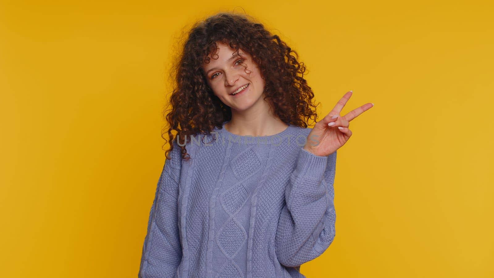 Happy curly haired young woman showing victory v sign, hoping for success and win, doing peace gesture, smiling with kind optimistic expression. Teen girl isolated alone on yellow studio background