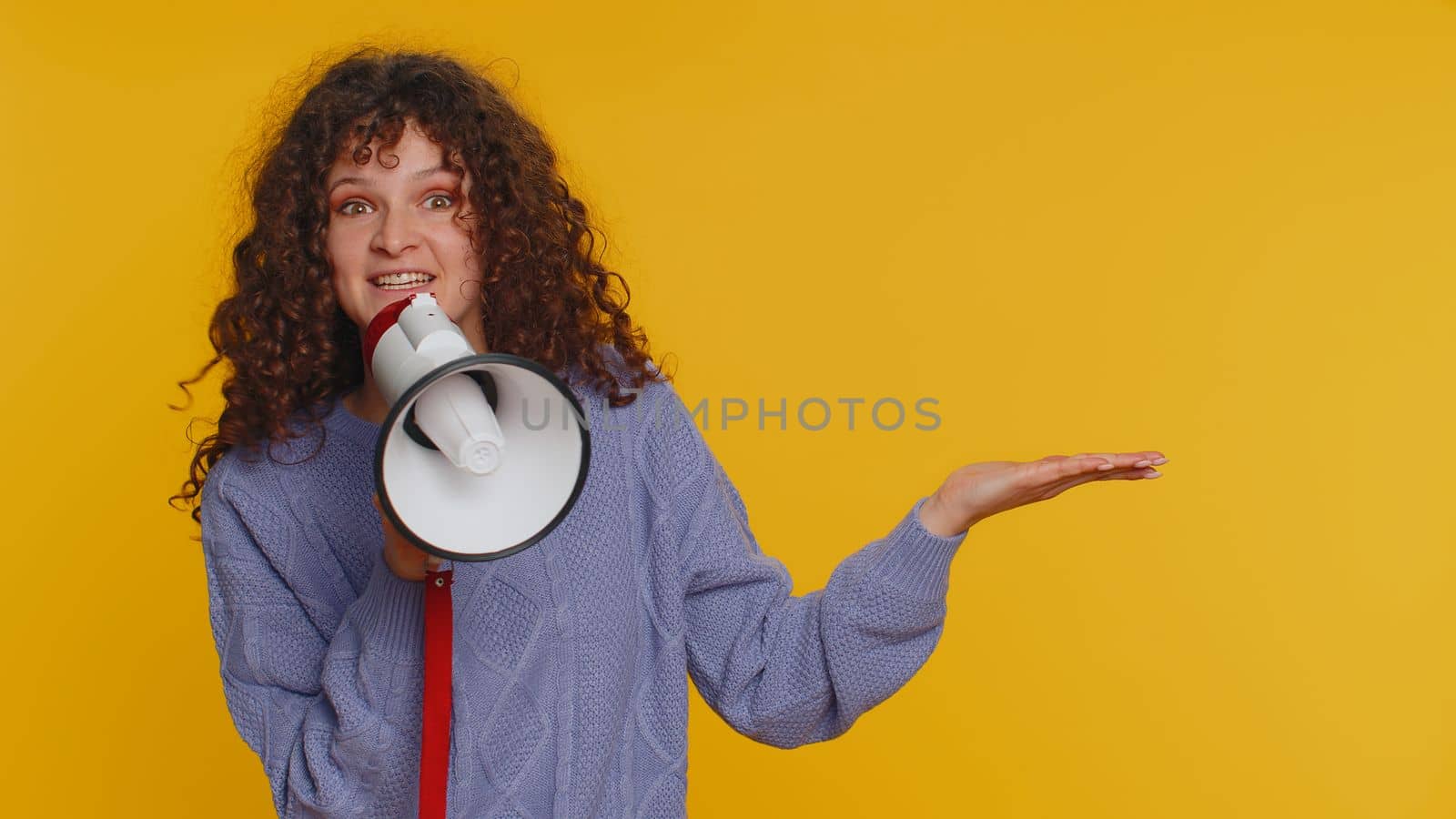 Happy woman talking with megaphone, proclaiming news, loudly announcing advertisement, warning using loudspeaker to shout speech, pointing empty place. Copy-space. Young girl on yellow background