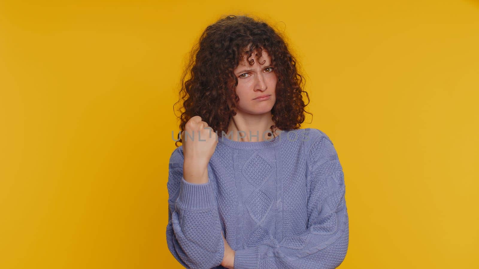 Aggressive angry curly haired woman trying to fight at camera, shaking fist, boxing with expression, punishment, disappointment, rage, quarrel. Young teen girl isolated on yellow studio background