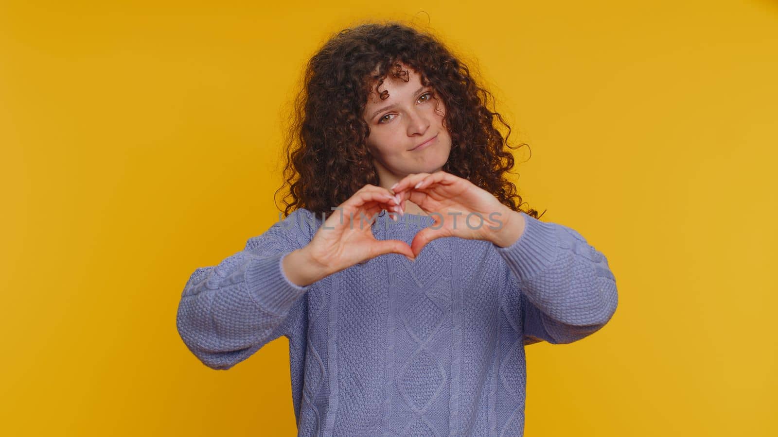 Woman in love. Smiling curly haired woman 20s in sweater makes heart gesture demonstrates love sign expresses good feelings and sympathy. Young pretty adult girl isolated on yellow studio background