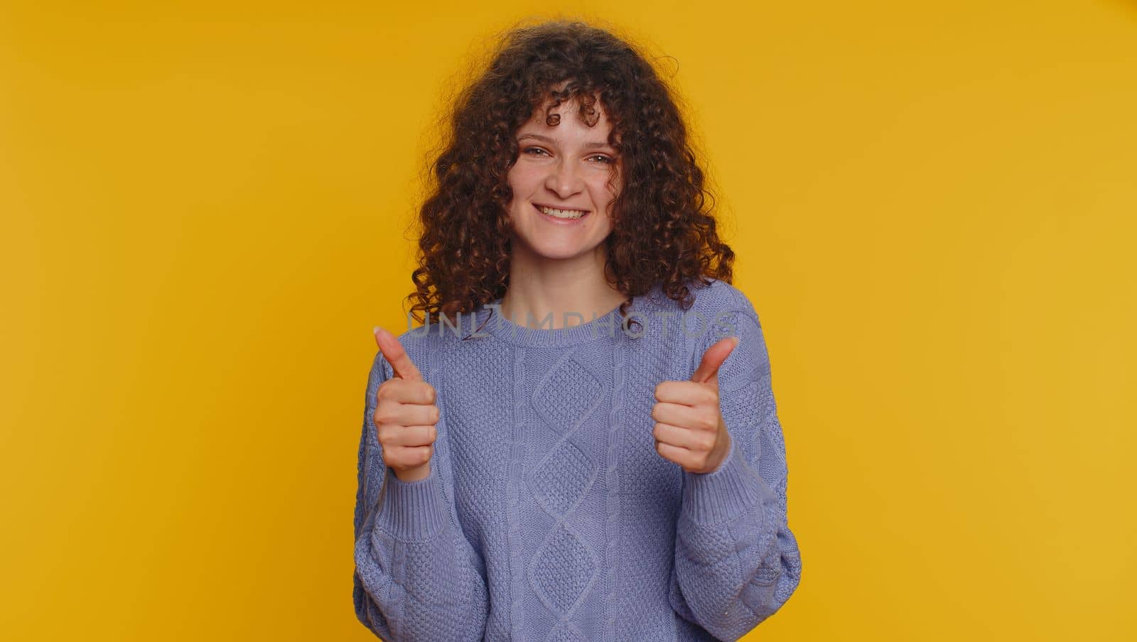 Like. Pretty curly haired woman in sweater raises thumbs up agrees with something or gives positive reply recommends advertisement likes good. Young girl isolated alone on yellow studio background