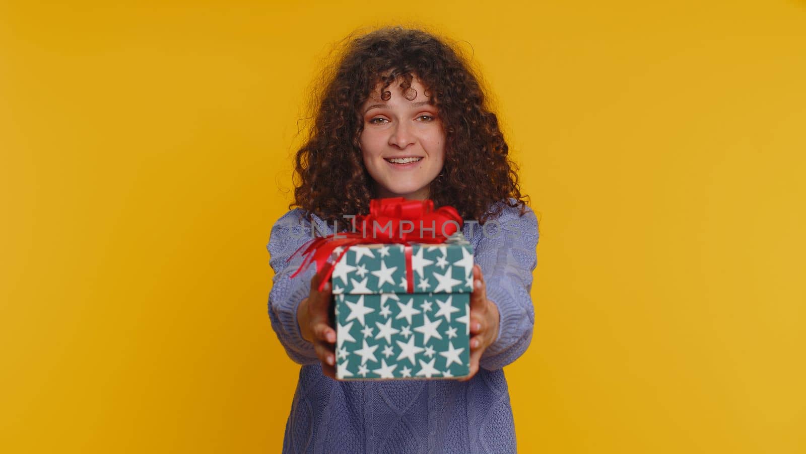 Positive smiling curly haired woman presenting birthday gift box stretches out hands, offer wrapped present career bonus, celebrating party. Young teen girl isolated alone on yellow studio background