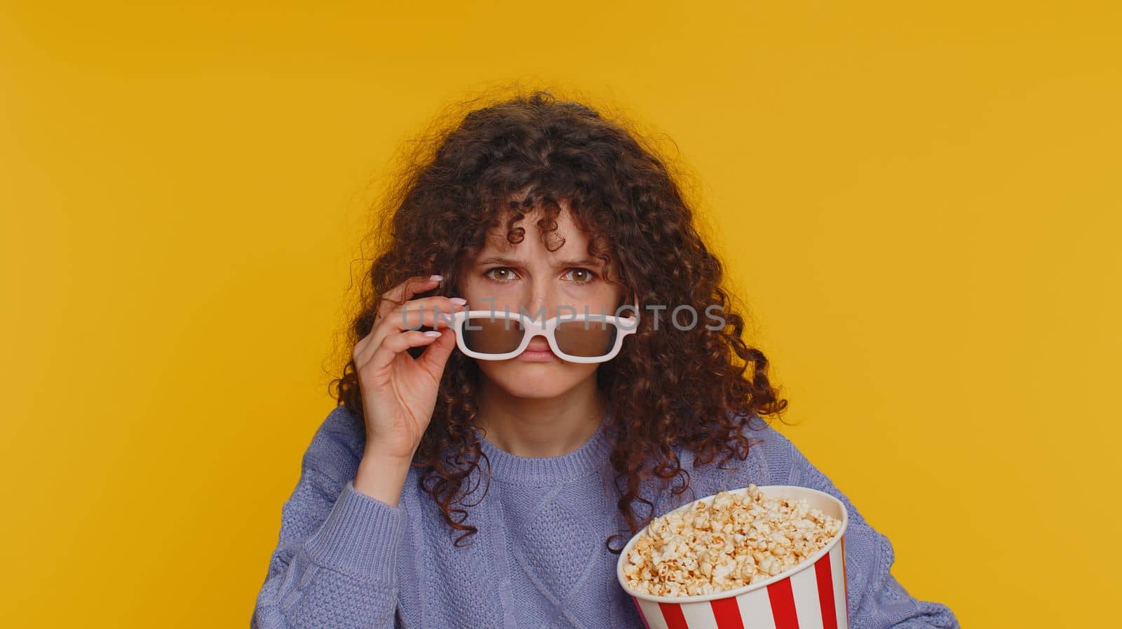 Excited curly haired woman in 3D glasses eating popcorn and watching interesting tv serial, sport game film online social media movie content. Girl enjoying domestic entertainment on yellow background