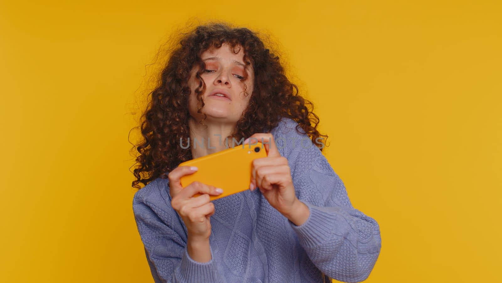 Worried funny addicted woman enthusiastically playing racing video games on mobile phone. Young curly haired girl using smartphone gadget app with drive simulator isolated on yellow studio background