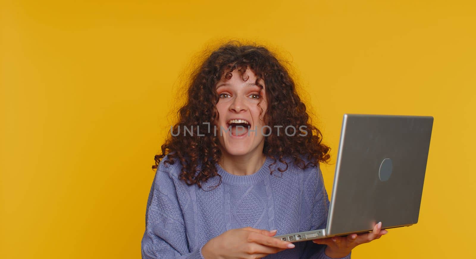 Overjoyed young curly haired woman use laptop scream in delight raise hands in triumph winner gesture celebrate success win money in lottery. Excited girl get online good news on yellow background