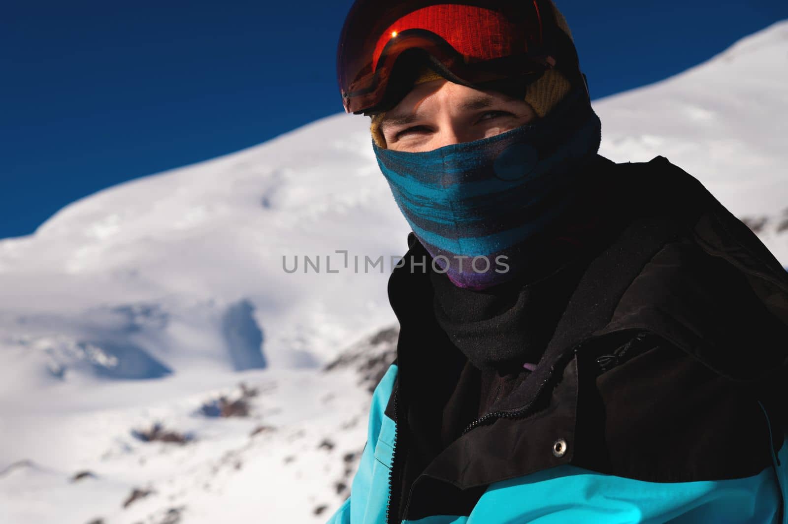 The skier looks at the camera before starting to ski. Man enjoys vacation in winter season. Portrait of a man in a balaclava and ski goggles against the backdrop of a snowy mountain by yanik88