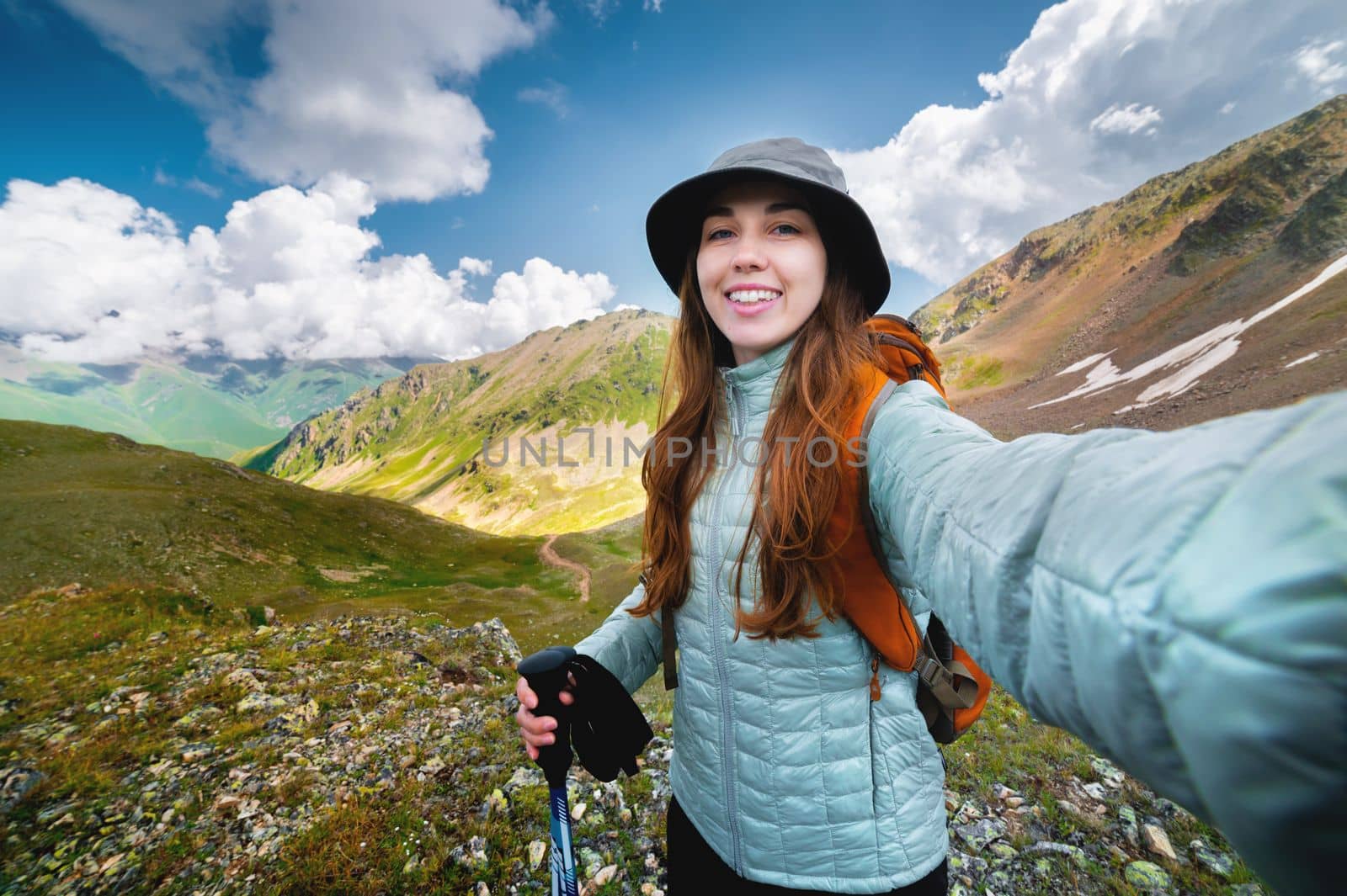 Smiling young woman takes a selfie on a mountain peak, with a backpack on a sunny day. Tourist hiking in the mountains.