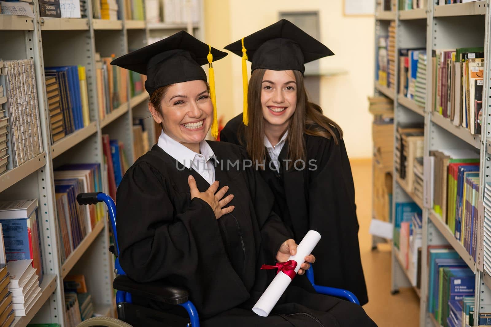 Happy young woman and woman in wheelchair in graduate gown with diploma in hands in library. Inclusive education. by mrwed54
