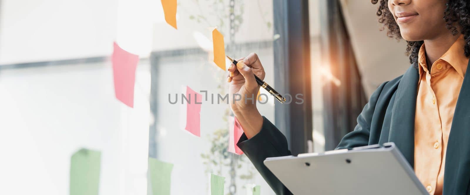 Attractive concentrated business lady in creating to-do list using multi coloured post-it sticky notes attaching them to transparent wall standing behind glass view, be more productive concept by wichayada