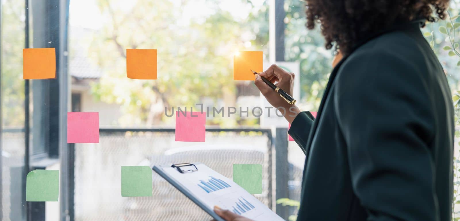 Attractive concentrated business lady in creating to-do list using multi coloured post-it sticky notes attaching them to transparent wall standing behind glass view, be more productive concept..