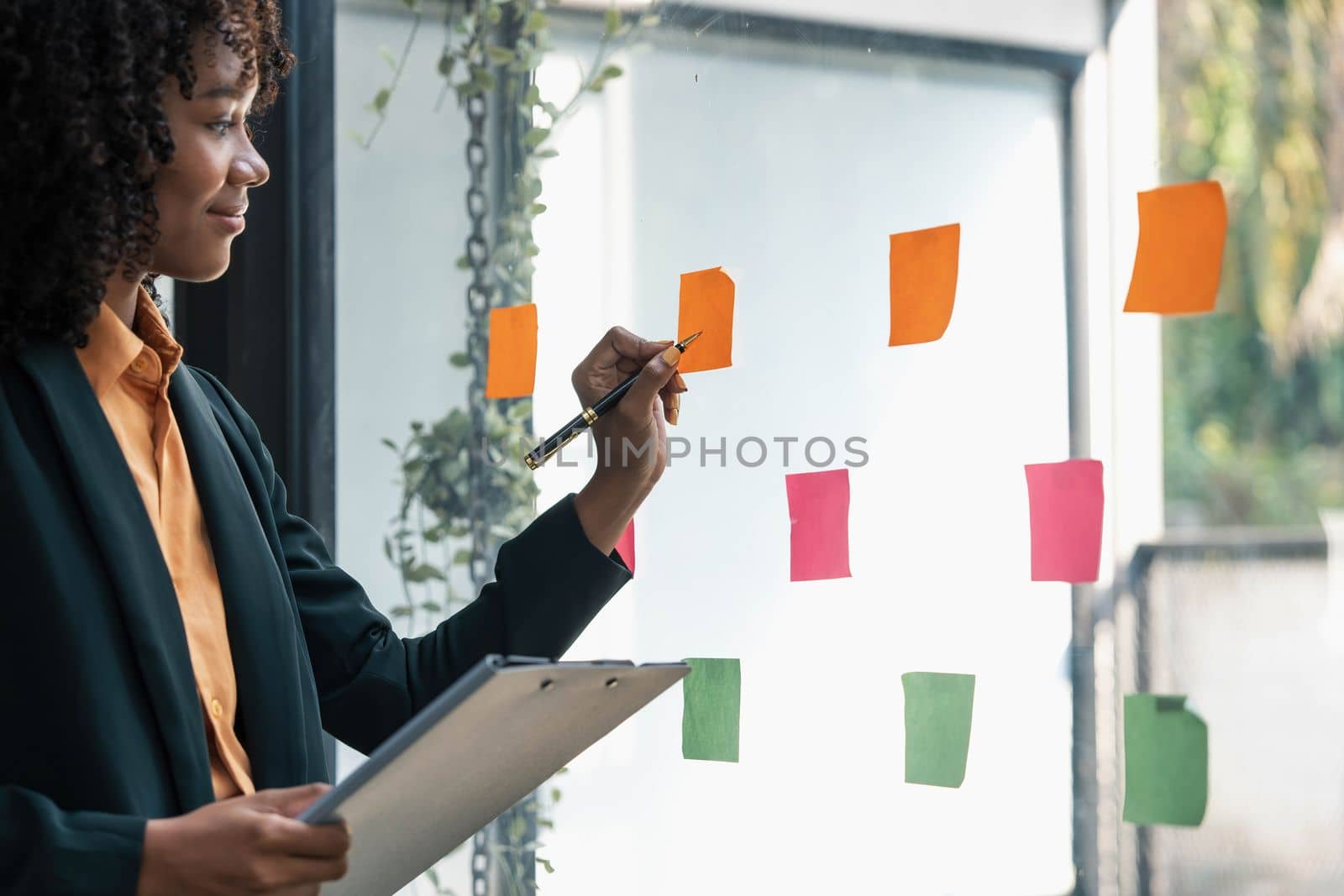 Attractive concentrated business lady in creating to-do list using multi coloured post-it sticky notes attaching them to transparent wall standing behind glass view, be more productive concept by wichayada