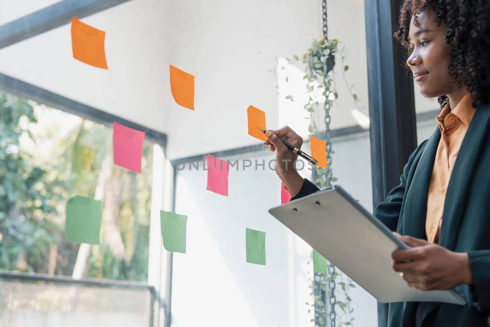 Attractive concentrated business lady in creating to-do list using multi coloured post-it sticky notes attaching them to transparent wall standing behind glass view, be more productive concept by wichayada