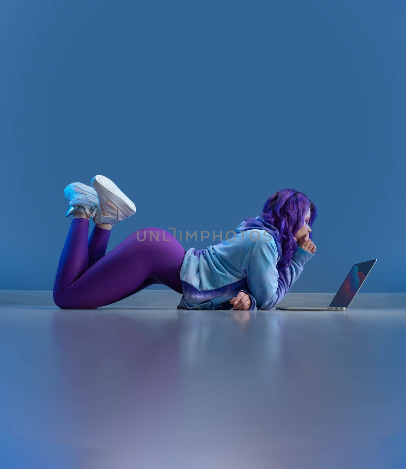 sexy girl in stylish sportswear and with purple hair sexy lying on the floor with a laptop on a blank blue background copy paste by Rotozey