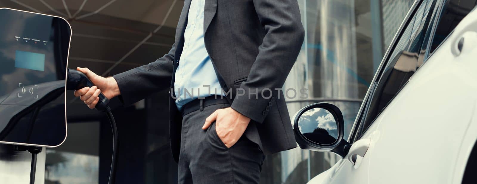 Closeup progressive businessman plugs charger plug from charging station to EV. by biancoblue