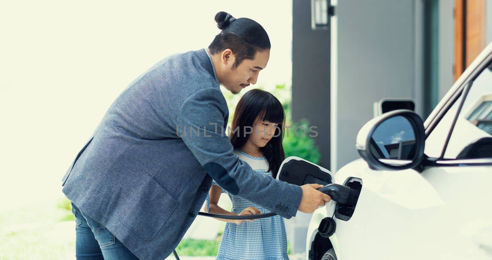 Progressive dad and daughter charging EV car from home charging station. by biancoblue