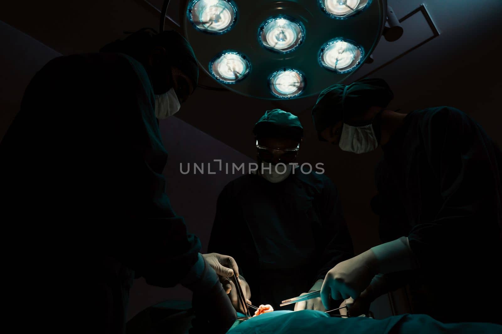 Surgical team performing surgery to patient in sterile operating room. In a surgery room lit by a lamp, a professional and confident surgical team provides medical care to an unconscious patient.