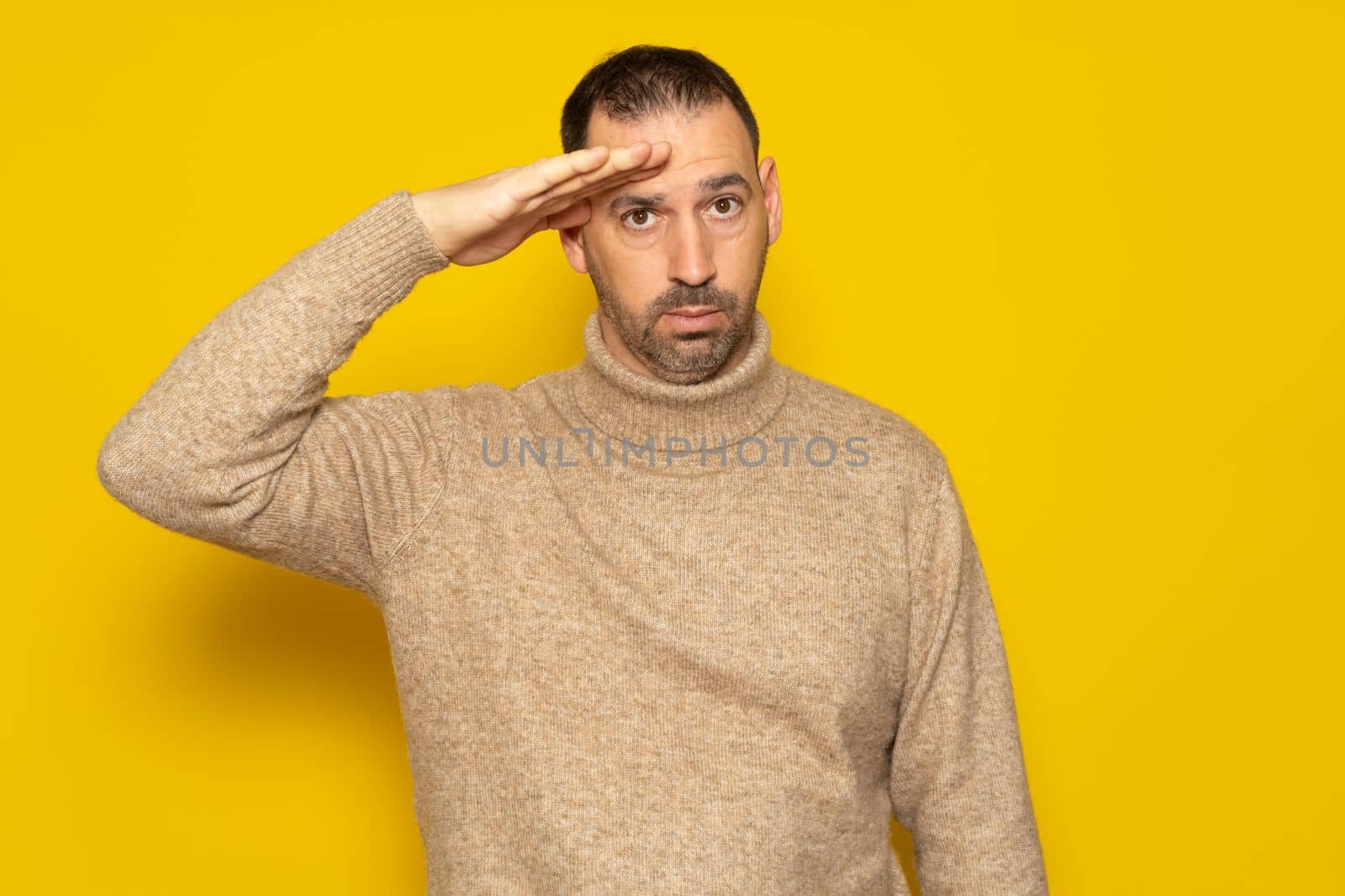 Handsome bearded Hispanic man wearing a turtleneck saluting the camera in an act of honor and patriotism, showing respect. Isolated on yellow background