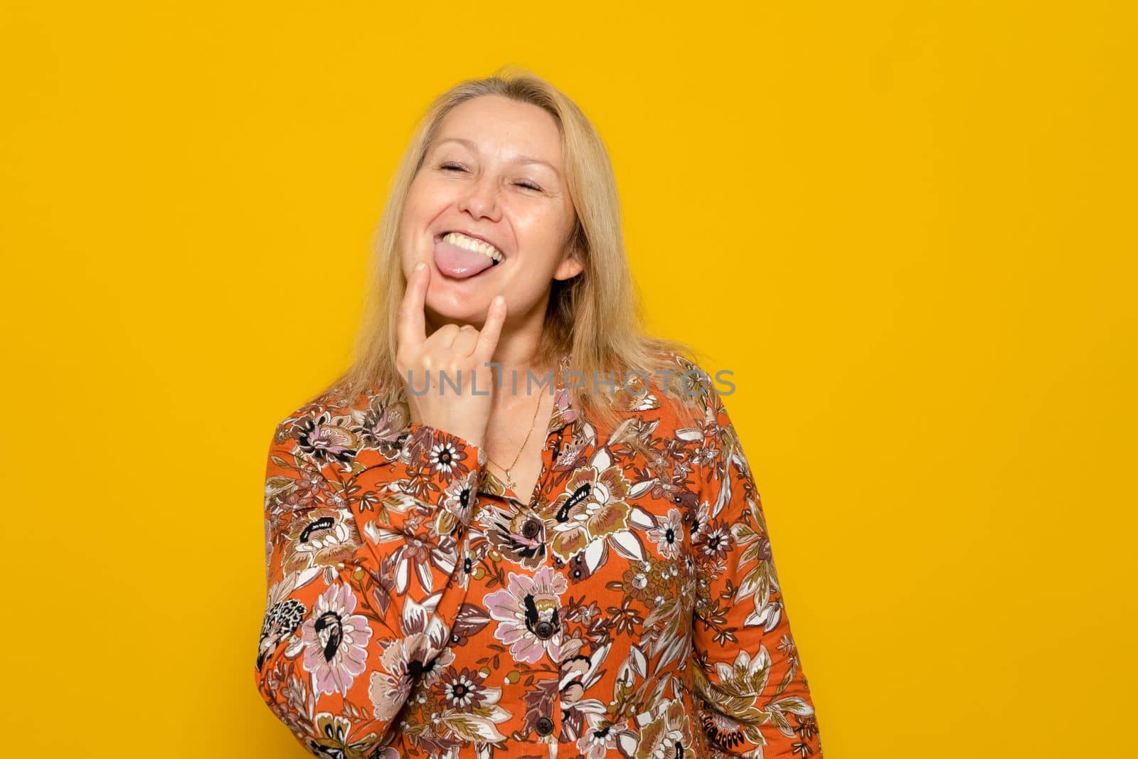 Caucasian blonde woman wearing a patterned dress making the rock gesture while sticking out her tongue in a carefree and cheeky attitude, isolated over yellow background