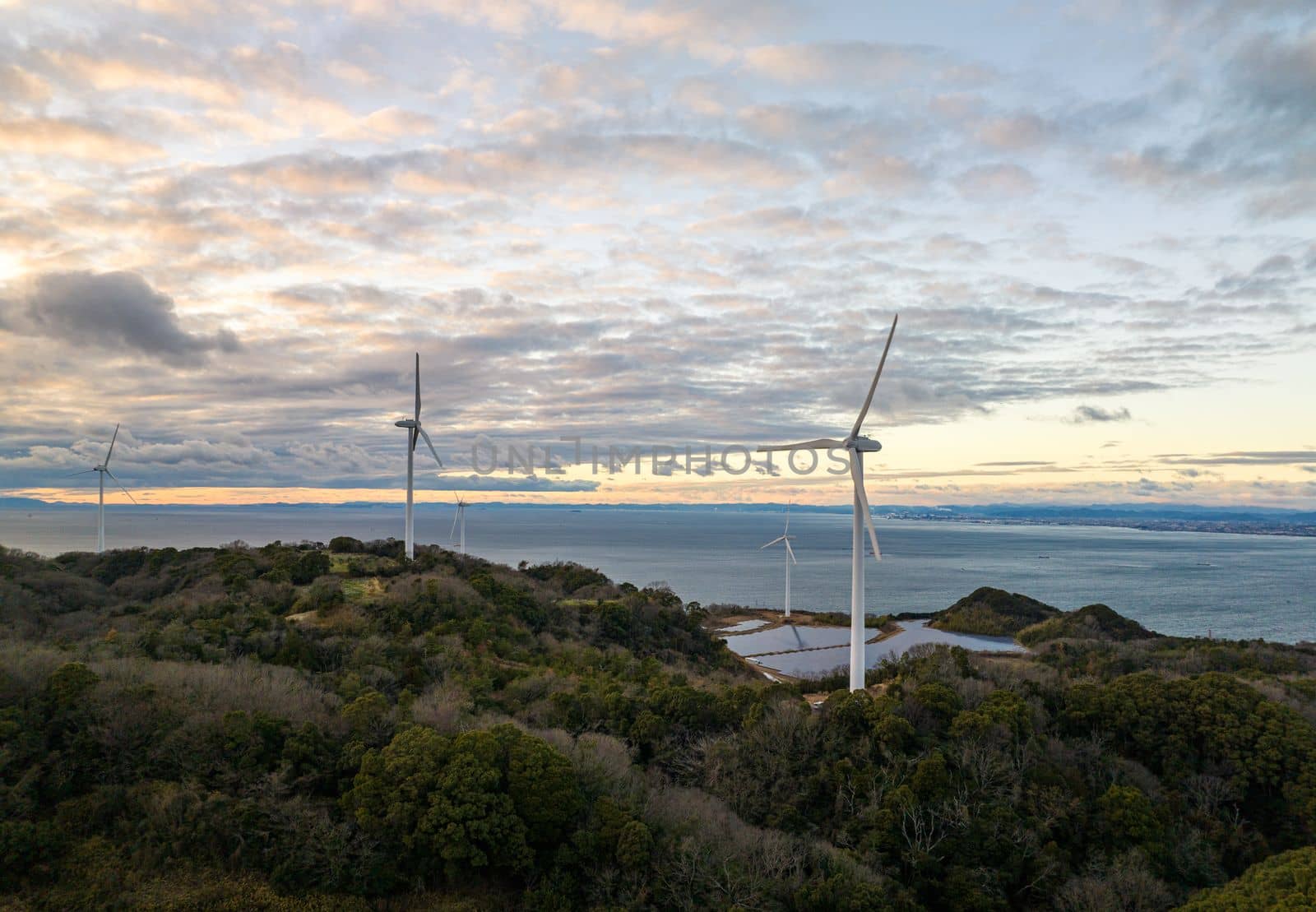 Wind Turbines Over Small Solar Farm in Green Landscape by Sea at Sunset by Osaze