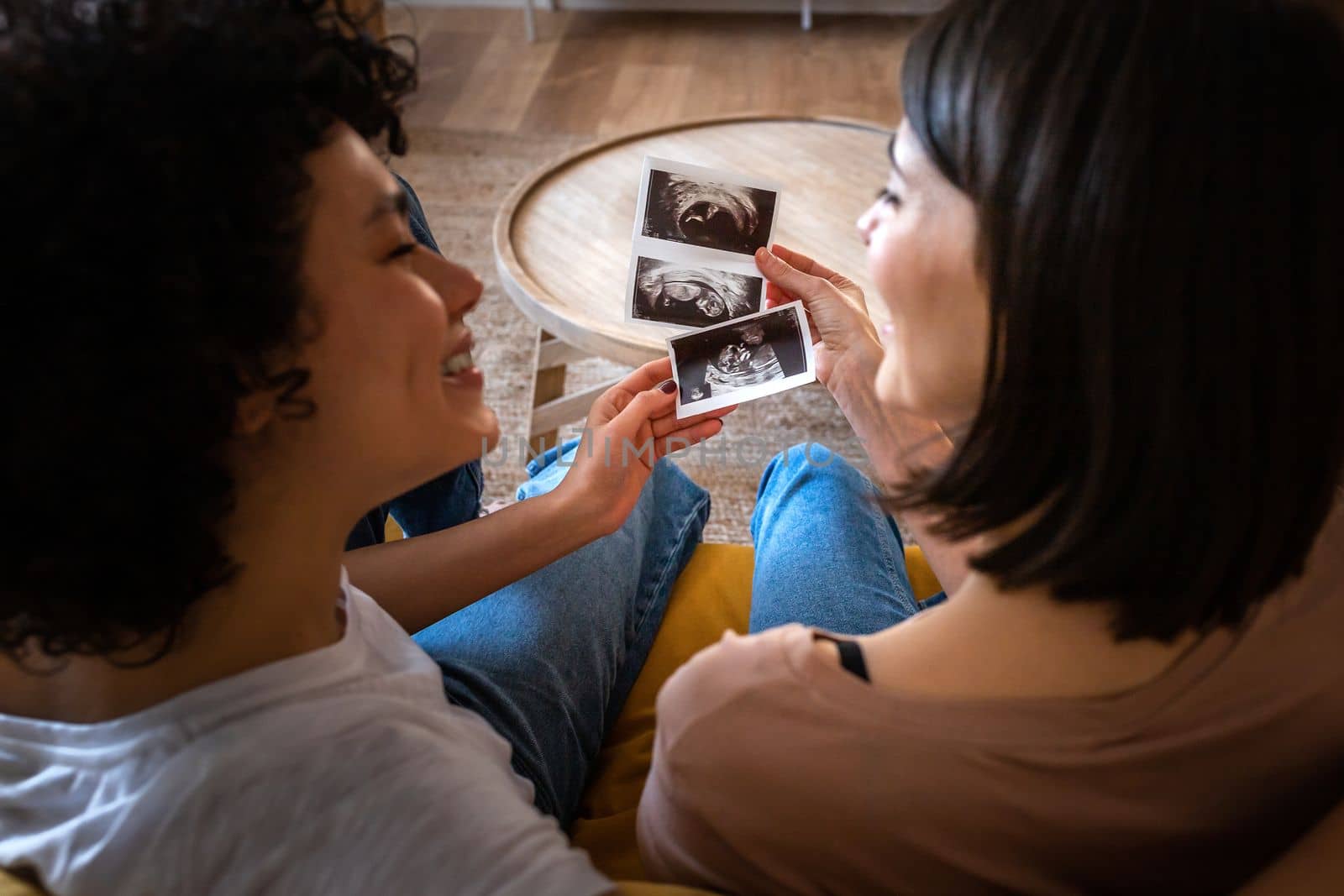 Top view of multiracial lesbian pregnant couple looking at each other in love while holding baby ultrasounds. LGTBQ love and family concepts.