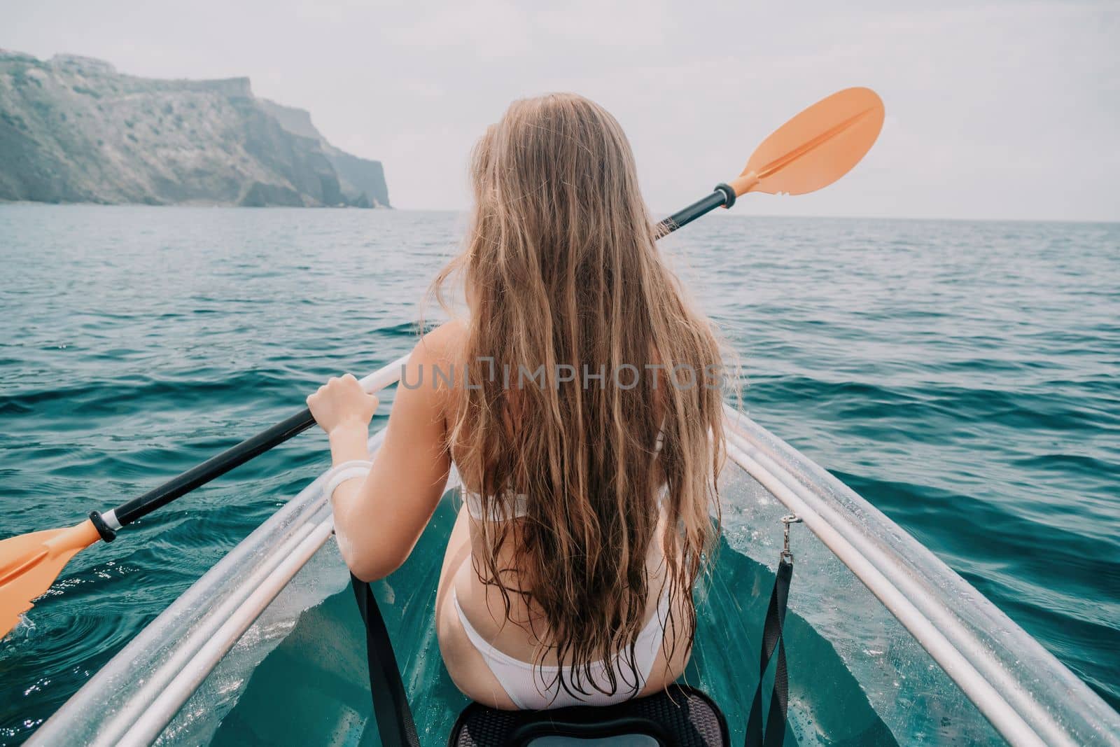 Woman in kayak back view. Happy young woman with long hair floating in transparent kayak on the crystal clear sea. Summer holiday vacation and cheerful female people having fun on the boat.